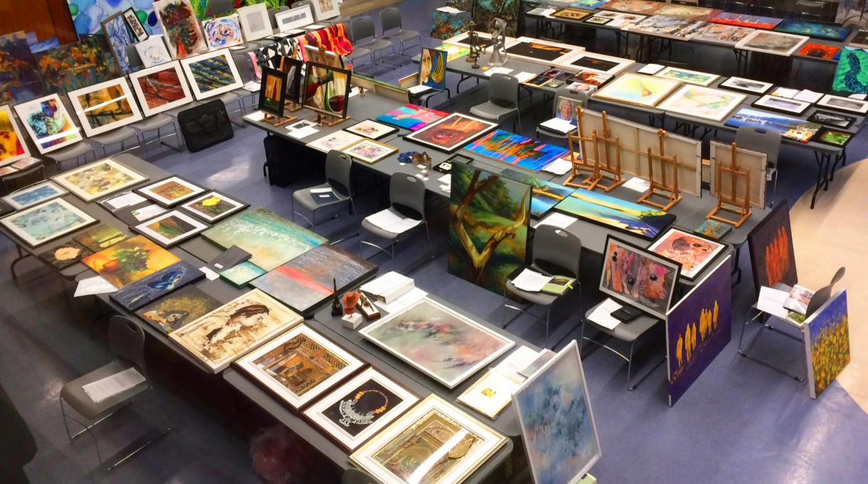 Large room with paintings laid out on tables and chairs.